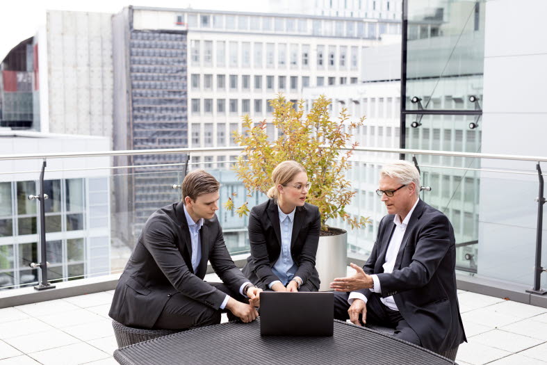 Bild på tre personer iklädda kostym som diskuterar framför en laptop.