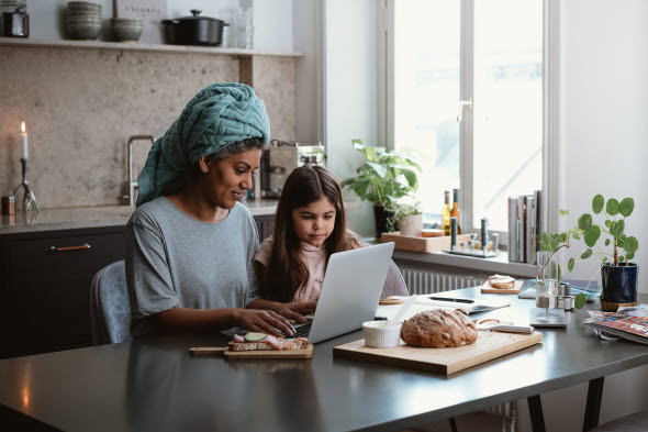 Mor och dotter som sitter i sitt kök och äter frukost samtidigt som de tittar på en laptop tillsammans