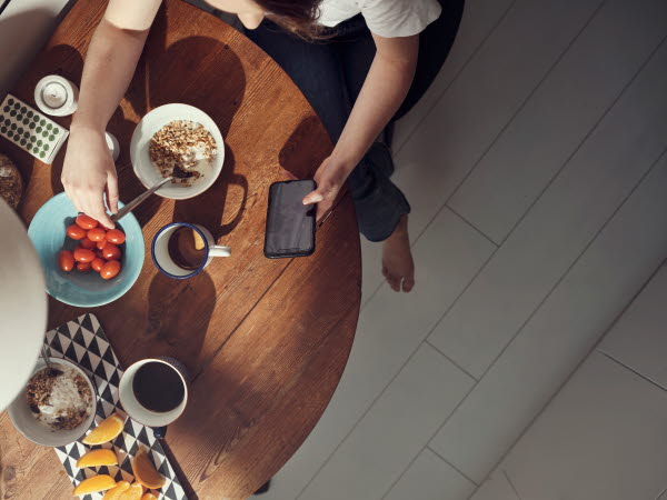 Bild på en person som sitter med sin mobiltelefon vid ett frukostbord.