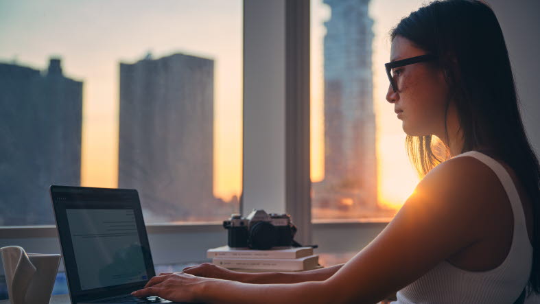 Ung kvinna jobbar vid en dator med utsikt över skyskrapor i bakgrunden.