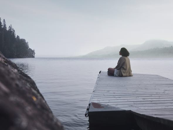 Kvinna sitter längst ut på en brygga och blickar ut över dimmig sjö.