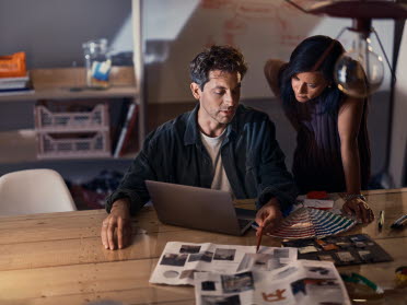 En man och en kvinna jobbar tillsammans i en studio vid ett skrivbord med tidningar utspridda på skrivbordet och med en laptop framför sig.