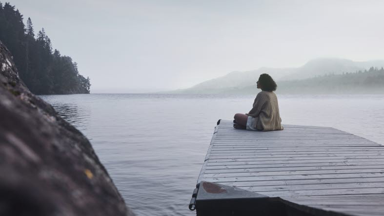 Kvinna sitter längst ut på en brygga och blickar ut över dimmig sjö.
