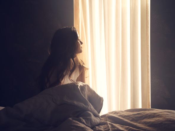 Kvinna som sitter på sin säng i sitt sovrum och tittar mot ett fönster
