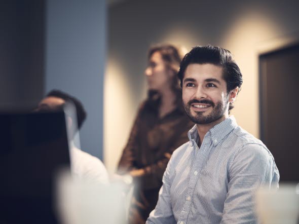 Man i skjorta och skägg ler mot sina kollegor i ett mötesrum på jobbet.
